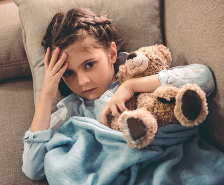 Sick little girl covered in blanket is hugging teddy bear and touching her forehead while lying on couchの写真素材