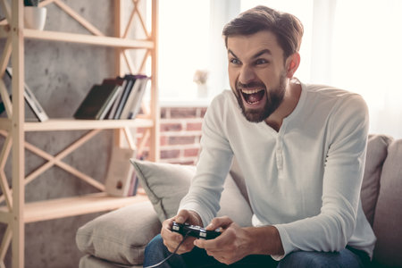 Handsome young man is playing game console and screaming while sitting on couch at home