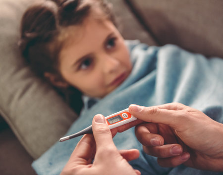 Sick little girl covered in blanket is lying on couch while her father is taking her temperature