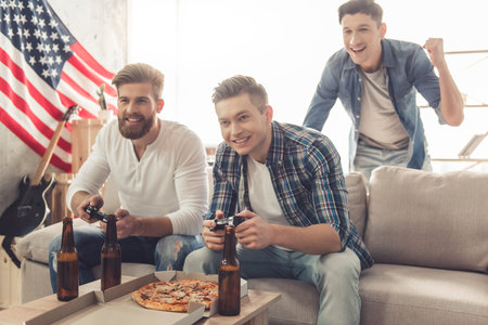 Attractive guys are playing video games, drinking beer, talking and smiling while sitting on couch...