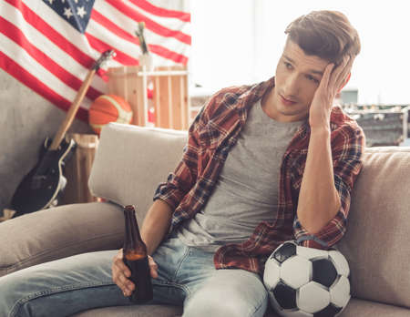 Attractive football fan is watching TV match and drinking beer while sitting on couch at homeの写真素材