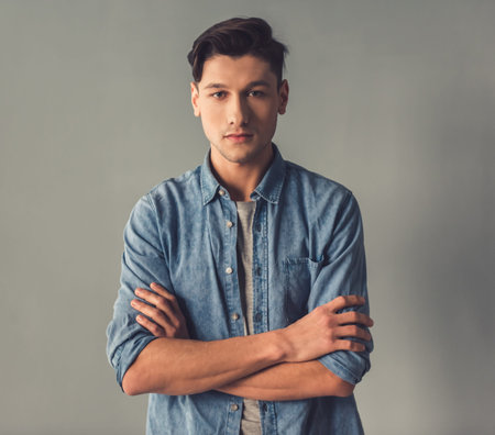 Attractive guy in casual clothes is looking at camera while standing with crossed arms on gray...