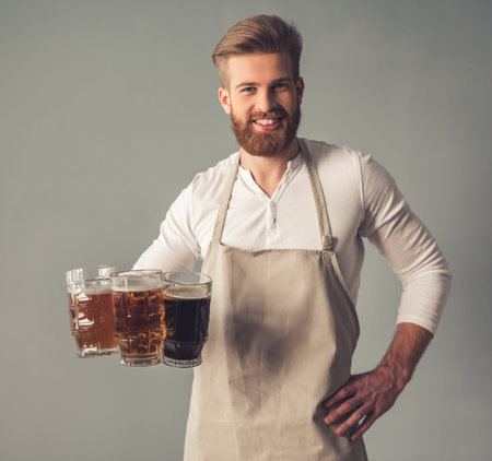 Handsome bearded waiter in apron is holding glasses of beer, looking at camera and smiling, on gray backgroundの写真素材