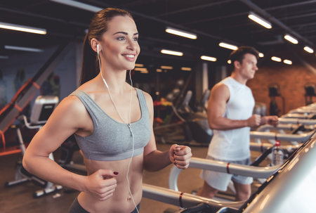 Attractive young woman in earphones is smiling while running on a treadmill in gym
