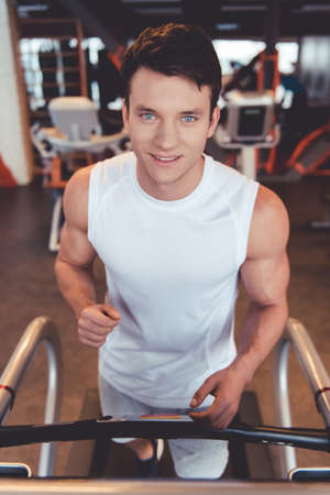 Attractive young muscular man is looking at camera and smiling while running on a treadmill in gymの写真素材