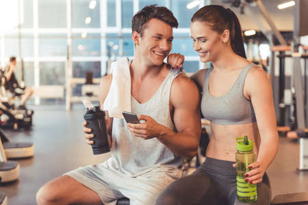Attractive young sports people are holding bottle of water, talking and smiling while resting in gymの写真素材