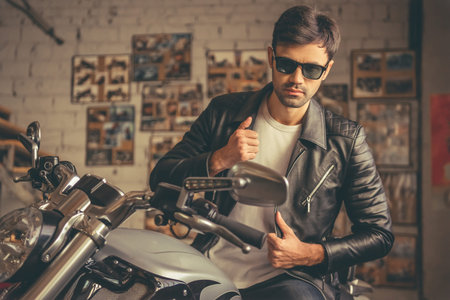 Attractive stylish guy in leather jacket and sun glasses is looking at camera while sitting on the motorcycle in the repair shopの写真素材
