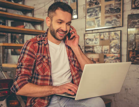 Handsome man in casual clothes is talking on the mobile phone, using a laptop and smiling while sitting in the motorcycle repair shopの写真素材