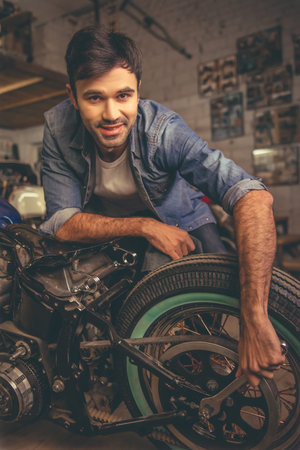 Attractive man is looking at camera and smiling while repairing a motorcycle in the repair shop
