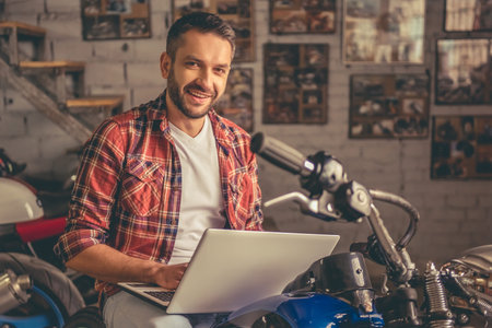 Handsome man in casual clothes is using a laptop, looking at camera and smiling while sitting on the...