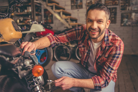 Handsome man is looking at camera and smiling while repairing a motorcycle in the repair shopの写真素材