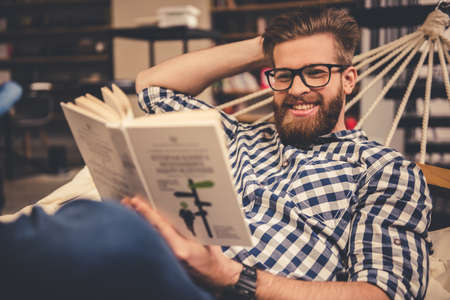 Handsome bearded young man is reading a book and smiling while lying in the hammock in modern libraryの写真素材