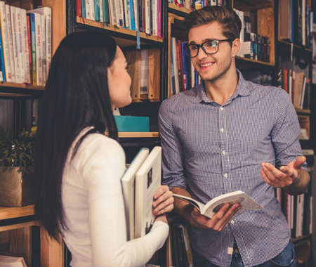 Beautiful young couple is talking and smiling while standing near the bookcase in the libraryの写真素材