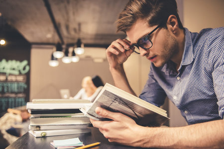 Concentrated young man is reading a book while working hard in the modern library