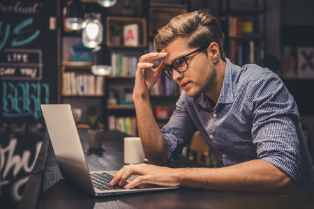 Attractive young man in eyeglasses is using a laptop while sitting in the modern library