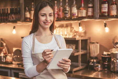 Beautiful female barista is making notes, looking at camera and smiling while standing near the bar counter in cafeの写真素材