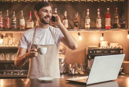 Handsome barista in apron is talking on the mobile phone, holding a cup of coffee and smiling while working at the bar counter in cafeの写真素材