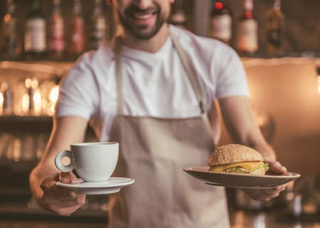 Handsome barista in apron is holding a cup of coffee and a sandwich and smiling while working at the bar counter in cafeの写真素材