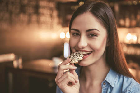 Beautiful girl is eating cookie, looking at camera and smiling while sitting at the cafeの写真素材
