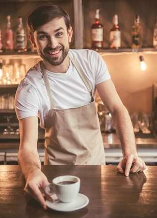Handsome barista in apron is holding a cup of coffee and smiling while working at the bar counter in cafeの写真素材