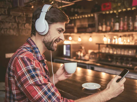 Handsome man in headphones is listening to music using a smartphone, drinking coffee and smiling while resting in cafeの写真素材
