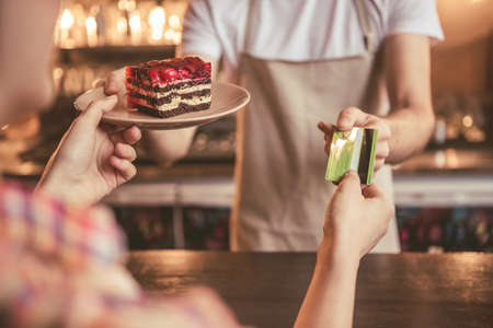 Cropped image of handsome barista in apron giving a cake and taking girl's credit card while working at the bar counter in cafeの写真素材