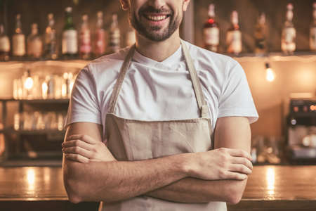 Cropped image of handsome barista in apron smiling while standing with crossed arms near the bar counter in cafeの写真素材