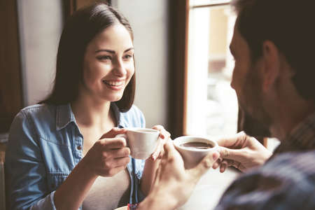 Happy young couple is drinking coffee, talking and smiling while sitting at the cafeの写真素材