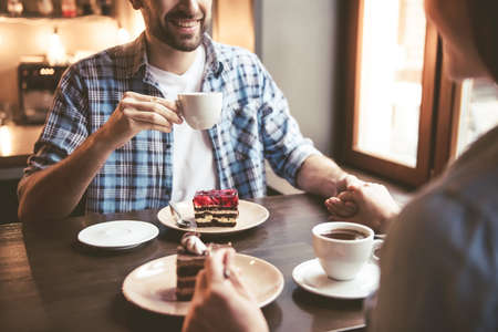 Cropped image of happy young couple holding hands, drinking coffee and smiling while sitting at the cafeの写真素材