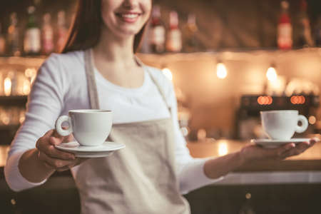 Cropped image of beautiful female barista holding a cups and smiling while standing near the bar counter in cafeの写真素材