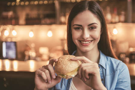 Beautiful girl is eating sandwich, looking at camera and smiling while sitting at the cafeの写真素材