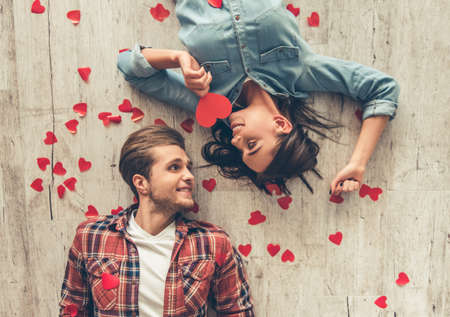 Top view of happy young couple looking at each other and smiling while lying on wooden floor. Girl is holding a red paper heartの写真素材
