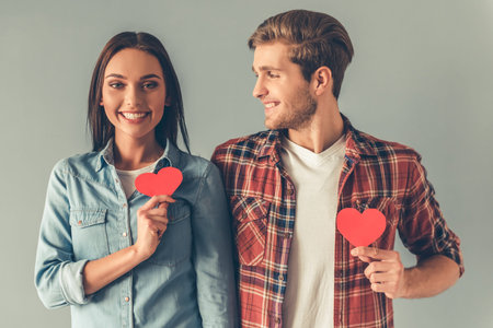 Happy young couple is holding red paper hearts and smiling, on gray background