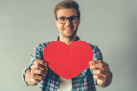 Handsome romantic guy is looking at camera and smiling while holding a red paper heart, on gray backgroundの写真素材