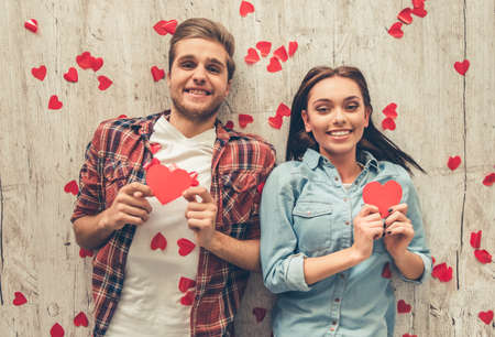 Top view of happy young couple holding red paper hearts, looking at camera and smiling while lying on wooden floorの写真素材