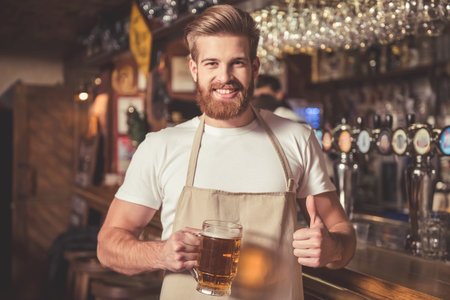 Handsome bearded bartender in apron is holding beer, showing Ok sign, looking at camera and smiling...