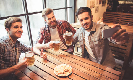 Handsome friends are doing selfie using a smart phone and smiling while drinking beer at the pub