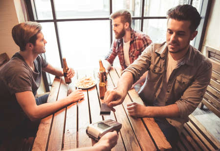 Handsome friends are drinking beer, talking and smiling while resting at the pub. One guy is paying using a credit cardの写真素材