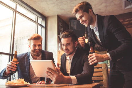 Handsome businessmen are drinking beer, using a digital tablet and smiling while resting at the pub
