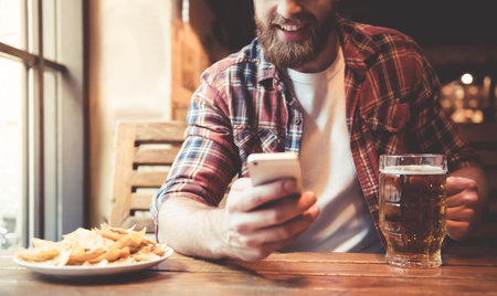 Cropped image of handsome bearded man using a smartphone and smiling while drinking beer in pubの写真素材