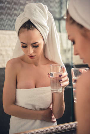 Beautiful girl in bath towel is feeling pain, holding a glass of water and looking into the mirror in bathroomの写真素材