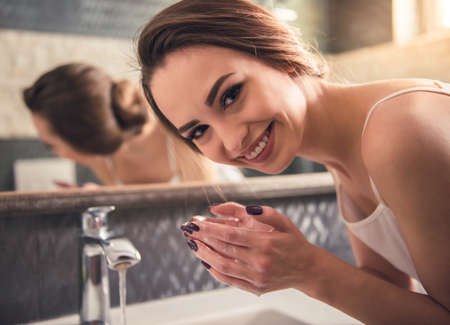 Beautiful girl in white undershirt is looking at camera and smiling while washing up in the bathroomの写真素材
