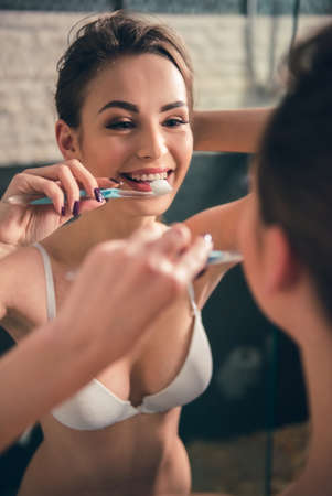 Beautiful girl in white bra is brushing teeth and smiling while looking into the mirror in bathroomの写真素材