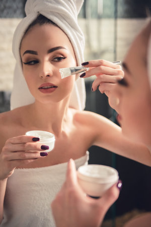 Beautiful girl in bath towel is applying cream on her face using a brush while looking into the mirror in bathroomの写真素材