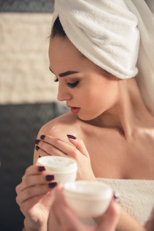Beautiful girl in bath towel is applying cream on her shoulder while standing in front of the mirror in bathroomの写真素材