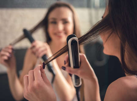 Beautiful girl in white undershirt is using a hair straightener and smiling while looking into the mirror in bathroomの写真素材