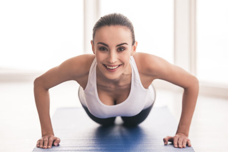 Beautiful girl in sportswear is looking at camera and smiling while doing push-ups on exercise mat