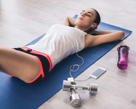 Beautiful girl in sportswear and earphones is listening to music while resting on yoga mat in sports hallの写真素材
