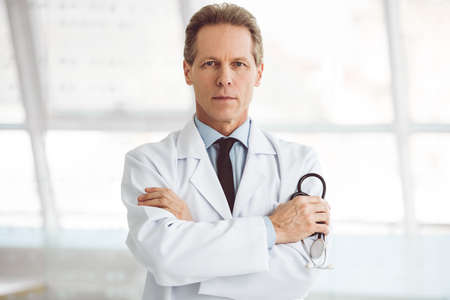 Handsome mature doctor is holding a stethoscope and looking at camera while standing with folded armsの写真素材