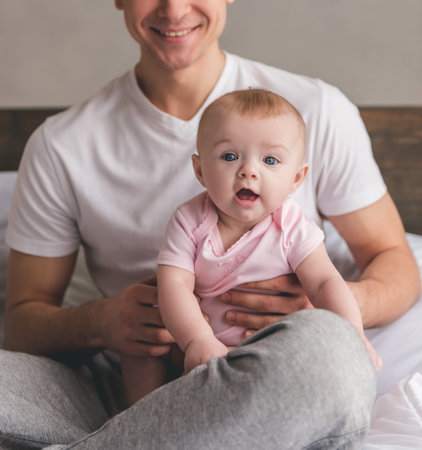 Handsome young dad is holding his cute baby in arms, looking at camera and smiling while sitting on bedの写真素材